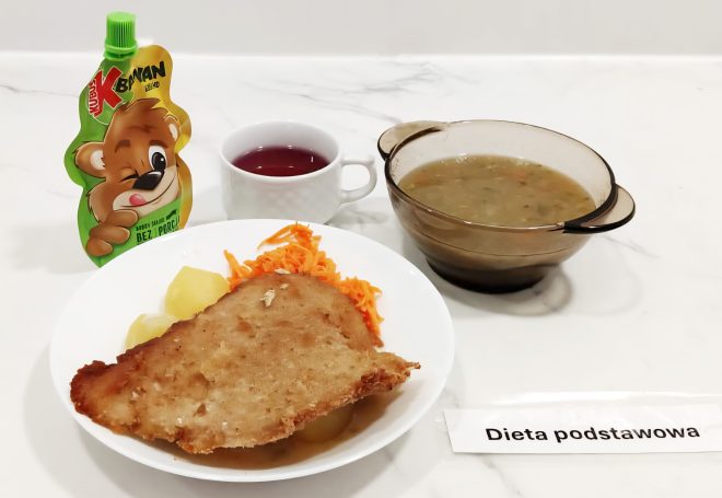 Na białym blacie ustawiony obiad w ramach diety podstawowej. Na talerzu panierowany kotlet schabowy, gotowane ziemniaki oraz surówka z marchwi. Obok kubek z czerwonym napojem, zupa jarzynowa w przezroczystej misce i deser w saszetce w kształcie misia z napisem "Banan". Obok kartka z napisem "Dieta podstawowa"