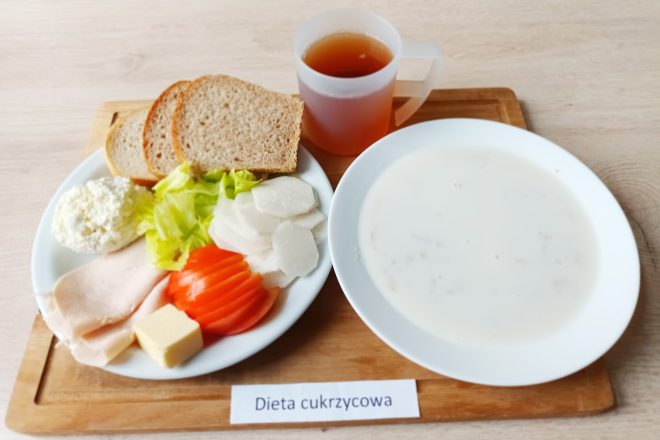 Taca z posiłkiem opisanym jako „Dieta cukrzycowa”. Na niej znajdują się dwa talerze oraz kubek z ciemnym napojem (prawdopodobnie herbata bez cukru). Na mniejszym talerzu: dwie kromki chleba razowego, plasterki pomidora i białej rzodkwi, liście sałaty, plasterki wędliny drobiowej, twarożek i kostka masła. Na większym talerzu – talerz mlecznej zupy, prawdopodobnie z płatkami zbożowymi