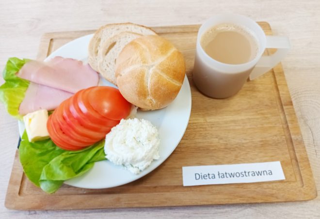 Śniadanie w diecie łatwostrawnej – na talerzu bułka pszenna, dwa plasterki chleba, szynka drobiowa, plasterki pomidora, liść sałaty, twarożek ziołowy i kawałek masła. Do tego kubek kawy z mlekiem. Wszystko podane na tacy z etykietą „Dieta łatwostrawna”