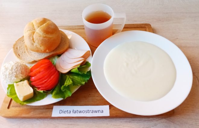 Na drewnianej tacy podano śniadanie składające się z dwóch kromek białego pieczywa i jednej bułki. Obok: plasterki chudej wędliny z indyka, plasterek pomidora, liść sałaty, twarożek oraz kawałek masła. Na oddzielnym talerzu kremowa zupa mleczna. Do picia – herbata w plastikowym kubku. Pod tacą widoczna etykieta z napisem „Dieta łatwostrawna”