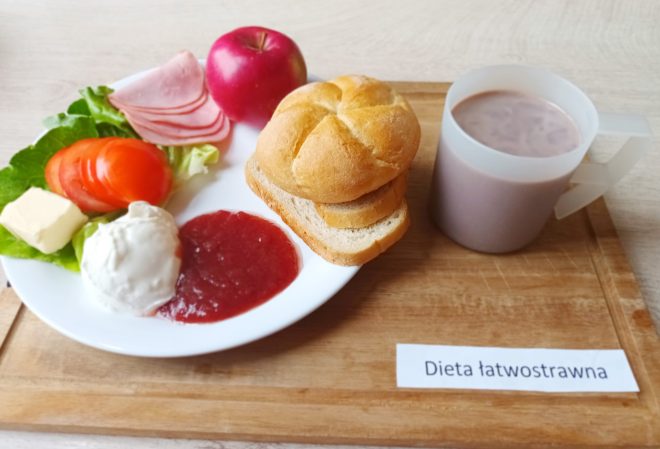 Talerz z posiłkiem w diecie łatwostrawnej: bułka pszenna i kromka chleba, plasterki szynki drobiowej, masło, pomidor, sałata, twarożek i galaretka owocowa. Obok świeże jabłko i kubek z napojem kakaowym. Wszystko ułożone na drewnianej tacy z etykietą "Dieta łatwostrawna"