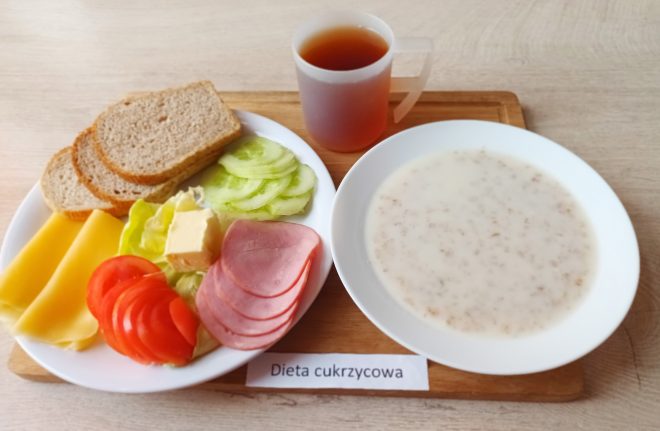 Pierwszy posiłek dnia to połączenie lekkostrawnych i wartościowych składników, dostosowanych do potrzeb osób z cukrzycą. Na talerzu znajdują się: pieczywo pełnoziarniste, plasterki żółtego sera i szynki drobiowej, świeże warzywa: pomidor, ogórek i sałata, porcja masła do pieczywa. Śniadanie uzupełnia pożywna owsianka na mleku, a całość dopełnia herbata bez cukru. To zbilansowane danie zapewnia stabilny poziom energii na dobry początek dnia i wspiera utrzymanie właściwego poziomu glukozy we krwi