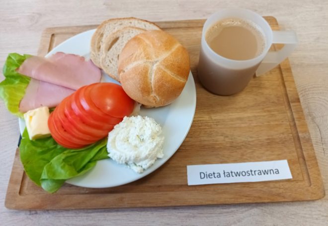 Taca z posiłkiem oznaczonym jako „Dieta łatwostrawna”. Na talerzu: bułka, dwa plasterki chleba, plastry szynki, plasterki pomidora, twarożek ze szczypiorkiem, listek sałaty, masło oraz kubek kawy z mlekiem