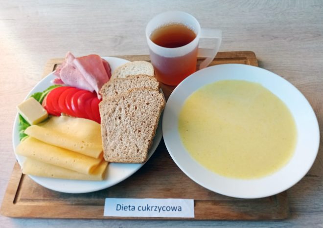 Zestaw śniadaniowy oznaczony jako „Dieta cukrzycowa” umieszczony na drewnianej tacy. Na talerzu znajduje się kilka kromek pieczywa mieszanego, plasterki żółtego sera, masło, plasterki pomidora, liść sałaty oraz dwa cienkie plastry wędliny. Obok talerza postawiono biały talerz z kremową zupą mleczną oraz przezroczysty kubek z herbatą