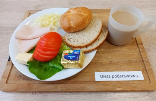 Na zdjęciu przedstawiono tacy z typowym śniadaniem szpitalnym w diecie podstawowej. Na białym talerzu leży kilka plasterków szynki drobiowej, porcja pokrojonego twarogu, plasterki pomidora i liść sałaty. Obok znajdują się również dwa kawałki pieczywa pszennego i bułka kajzerka, a także kostka sera żółtego Gouda w opakowaniu oraz kawałek masła. Do posiłku podany jest jasnobeżowy napój w plastikowym kubku z uchem – może to być kawa z mlekiem lub kawa zbożowa. Zestaw śniadaniowy umieszczony jest na drewnianej tacy z podpisem „Dieta podstawowa”