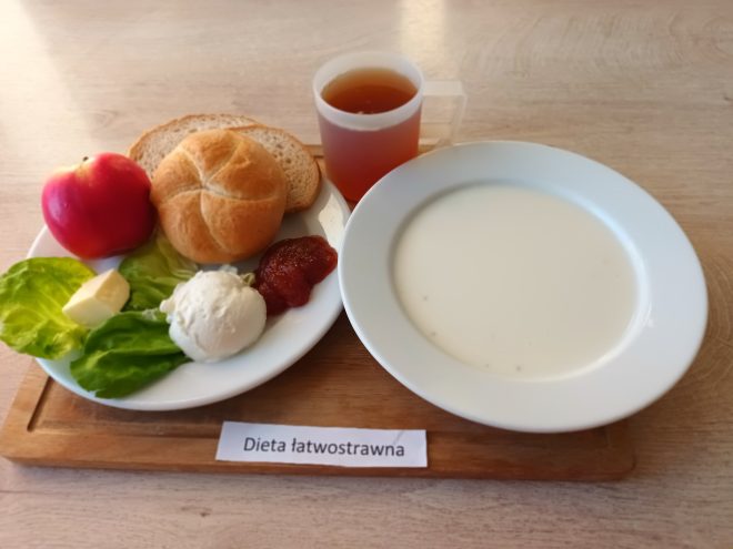 Zestaw śniadaniowy w diecie łatwostrawnej. Na tacy znajduje się talerz z białą zupą mleczną, a obok drugi talerz z pieczywem pszennym (bułką i kromką chleba), twarożkiem, liściem sałaty z masłem, musem owocowym i czerwonym jabłkiem. Do posiłku podano herbatę w przezroczystym kubku z rączką. Taca oznaczona jest napisem „Dieta łatwostrawna”