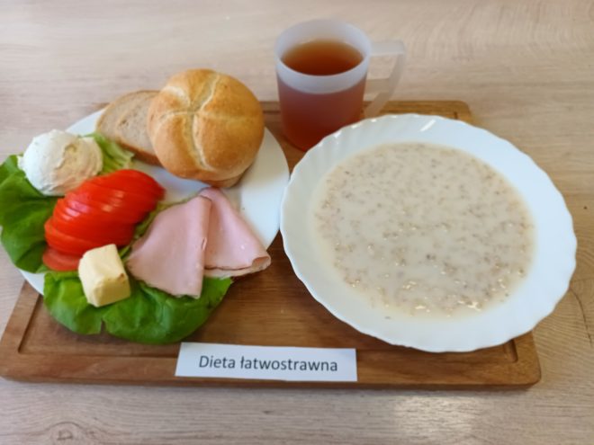 Śniadanie w wersji łatwostrawnej. Na drewnianej tacy znajdują się dwa talerze: jeden z owsianką mleczną, drugi z bułką pszenną, kromką chleba, liściem sałaty, plasterkami pomidora, twarożkiem, plastrem szynki drobiowej oraz masłem. Obok umieszczony jest kubek z herbatą. Dieta oznaczona karteczką z napisem „Dieta łatwostrawna”