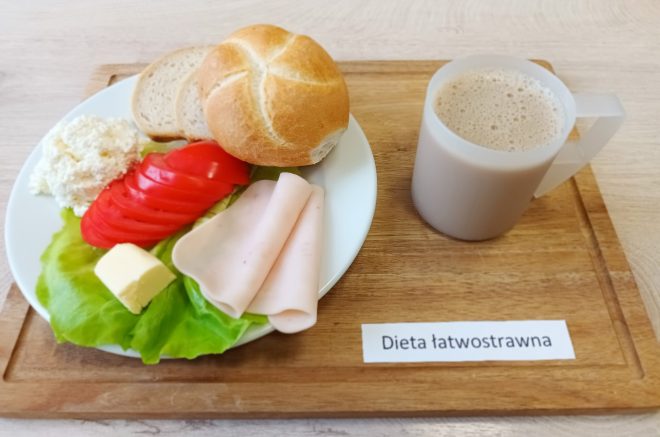Drewniana taca z talerzem zawierającym bułkę kajzerkę, dwa plasterki chleba, twarożek, masło, pomidora w plastrach, szynkę drobiową i liść sałaty. Obok kubek z kawą z mlekiem. Karteczka z napisem „Dieta łatwostrawna”