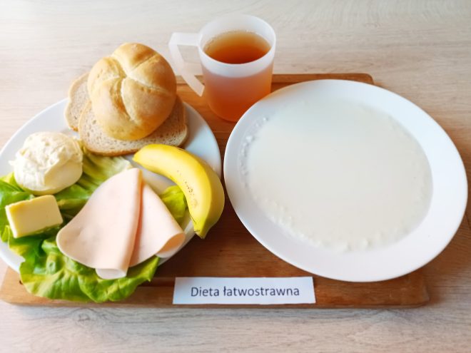 Śniadanie w diecie łatwostrawnej: na talerzu bułka kajzerka, dwie kromki chleba pszennego, plaster szynki drobiowej, kawałek masła, twarożek, liść sałaty i banan. Obok talerz z mleczną kaszką (lub kleikiem ryżowym) oraz kubek z herbatą. Całość podana na tacy z podpisem „Dieta łatwostrawna”