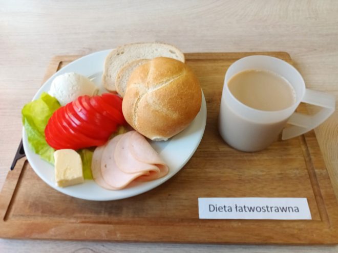 Śniadanie w diecie łatwostrawnej. Na talerzu znajduje się bułka kajzerka, dwie kromki chleba pszennego, plasterki wędliny drobiowej, pokrojony pomidor, liść sałaty, serek twarogowy oraz kostka masła. Obok przezroczysty kubek z kawą z mlekiem