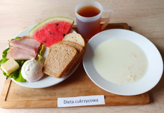 Śniadanie w diecie cukrzycowej podane na drewnianej tacy. Na jednym talerzu znajdują się dwa rodzaje pieczywa – kromki chleba razowego i jasnego, plasterki wędliny, kostka masła, kulka twarożku oraz duży trójkątny kawałek arbuza, wszystko ułożone na liściu sałaty. Obok miska z jasną, mleczną zupą z drobną kaszką. W górnym rogu przezroczysty kubek z herbatą. Na tacy widoczna etykieta z napisem „Dieta cukrzycowa”