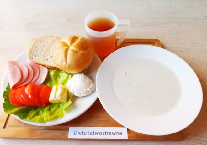 Śniadanie szpitalne w wersji łatwostrawnej. Na talerzu leżą dwie kromki chleba pszennego i bułka kajzerka, plasterki drobiowej wędliny, masło, serek twarogowy, liść sałaty, plasterki świeżych pomidorów i papryki. Obok znajduje się talerz z jasną, mleczną zupą oraz kubek z herbatą owocową. Na tacy widnieje etykieta z napisem „Dieta łatwostrawna”