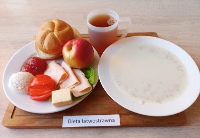 Taca z posiłkiem śniadaniowym oznaczonym jako „Dieta łatwostrawna”. Na białym talerzu znajdują się dwie bułki pszenne, plasterki chudej wędliny z indyka, pomidor, twarożek, dżem truskawkowy, kawałek masła, liść sałaty i jabłko. Obok talerza znajduje się przezroczysty kubek z herbatą oraz talerz z mleczną zupą na bazie kaszy manny lub ryżu