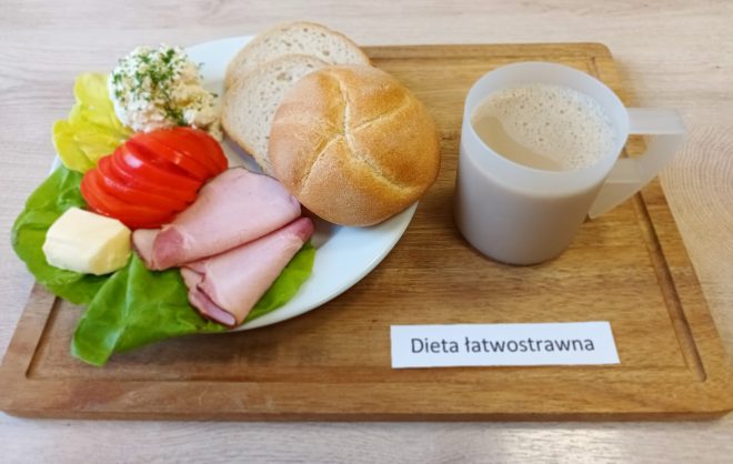 Talerz z bułką pszenną i dwoma kromkami chleba pszennego. Obok porcja szynki gotowanej, masła, plasterki pomidora i sałata. Dodatkowo twarożek z koperkiem. Do posiłku dołączono kawę z mlekiem w plastikowym kubku. Posiłek w ramach diety łatwostrawnej