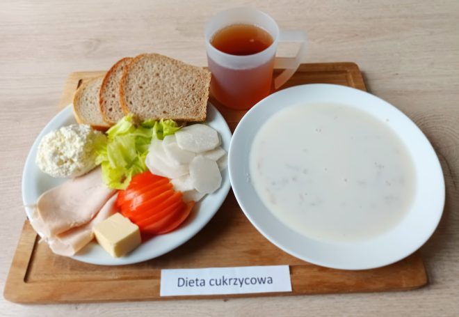 Białe pieczywo pszenne w towarzystwie gotowanego jajka, plasterka szynki, ogórka konserwowego, liścia sałaty i czerwonej papryki. Na talerzu również porcja masła. Obok kubek z kawą z mlekiem. Danie podane na białym talerzu i podpisane jako „Dieta podstawowa”