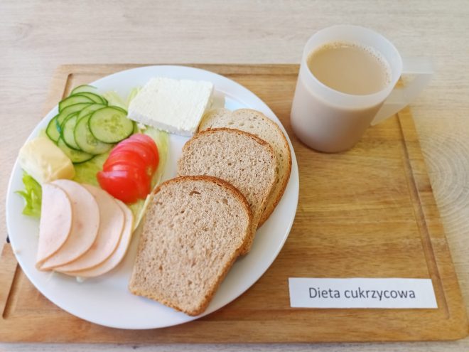 Taca z posiłkiem dla osoby na diecie cukrzycowej. Na białym talerzu znajdują się trzy kromki chleba razowego, plasterki wędliny drobiowej, plasterki ogórka i pomidora, plaster sera białego oraz liść sałaty i kawałek masła. Obok na tacy stoi przezroczysty kubek z kawą z mlekiem. Taca jest podpisana etykietą: „Dieta cukrzycowa”