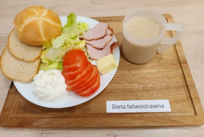 Taca z lekkostrawnym śniadaniem. Na talerzu: bułka kajzerka, dwa plasterki chleba pszennego, kilka plasterków pomidora, liście sałaty, plasterki chudej wędliny, porcja białego serka i kostka masła. Obok przezroczysty kubek z kawą z mlekiem lub zbożówką