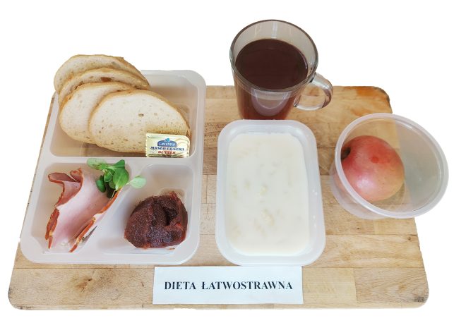 Taca z posiłkiem oznaczonym jako „Dieta łatwostrawna”. W zestawie znajdują się cztery kromki chleba pszennego, plasterki szynki, liść zieleniny, porcja ciemnej konfitury, kostka masła w opakowaniu marki Lactima, pojemnik z mleczną kaszką lub zupą, jabłko w plastikowym pudełku oraz szklanka ciemnej herbaty