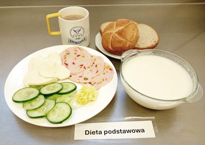 Taca z zestawem śniadaniowym diety podstawowej. Na białym talerzu plasterki wędliny drobiowej z warzywami, plastry białego sera, plasterki świeżego ogórka, listki sałaty oraz porcja masła. Obok talerza miseczka z mlekiem lub zupą mleczną. W tle bułka kajzerka i kromki chleba pszennego na osobnym spodku oraz kubek z kawą zbożową w białym kubku z żółtym uchem, opatrzonym logotypem „Kryniczanka”. Wszystko ustawione na metalowym blacie z etykietą „Dieta podstawowa”