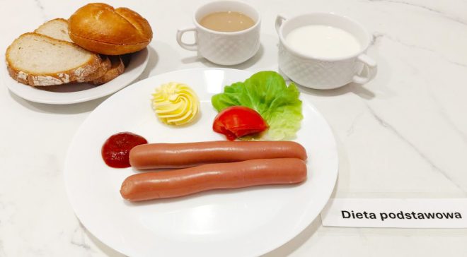 Talerz śniadaniowy z dwoma parówkami, porcją ketchupu, masłem dekoracyjnie uformowanym, liściem sałaty i plastrem pomidora. Obok znajdują się filiżanka z kawą z mlekiem, filiżanka z białą kawą zbożową, miseczka z mlekiem oraz talerzyk z pieczywem – bułką i kromkami chleba pszennego. Całość opatrzona karteczką "Dieta podstawowa"