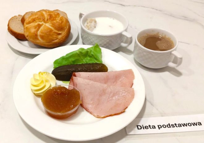 Taca z posiłkiem śniadaniowym oznaczonym jako „Dieta podstawowa”. Na talerzu dwie plastry szynki, sałata, ogórek kiszony, porcja masła i galaretka jabłkowa w pojemniczku. Obok bułka i kromki chleba pszennego na osobnym talerzyku. W zestawie również kawa z mlekiem oraz miseczka z mlekiem i płatkami