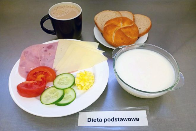 Taca z posiłkiem oznaczonym jako „Dieta podstawowa”. Na talerzu plastry szynki i sera żółtego, ogórek, pomidor, porcja masła. Obok bułka i kromki chleba pszennego oraz czarna filiżanka z kawą z mlekiem. Dodatkowo miseczka z białym jogurtem lub kefirem