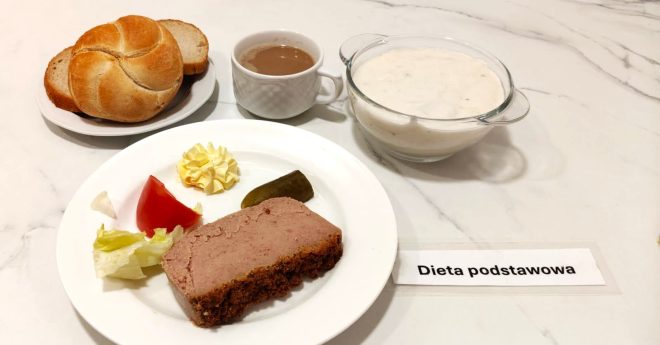 Śniadanie w ramach diety podstawowej: porcja pasztetu z ogórkiem konserwowym, masło, kawałek pomidora i sałata lodowa na talerzu. Obok filiżanka kawy z mlekiem, miseczka białej zupy mlecznej oraz bułka kajzerka i kromka chleba. Wszystko ułożone na jasnym blacie z etykietą „Dieta podstawowa”