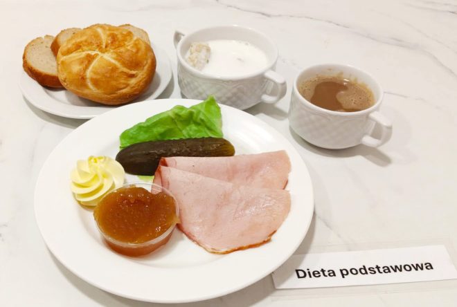 Talerz z dwoma plastrami szynki, sałatą, ogórkiem konserwowym, porcją masła i galaretki owocowej. Obok bułka kajzerka i kromki chleba na talerzyku, filiżanka z kawą i miseczka z białym twarożkiem zalanym mlekiem. Karteczka z napisem „Dieta podstawowa”