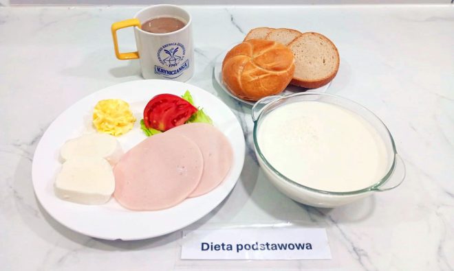 Rozbudowane śniadanie w diecie podstawowej. Na talerzu dwa plasterki wędliny drobiowej, dwa plasterki białego sera, porcja masła, plaster pomidora i sałata. Obok kubek z kawą z mlekiem z logo „Kryniczanka”, miska z białym napojem mlecznym (prawdopodobnie kefirem lub mlekiem), bułka kajzerka i trzy kromki chleba pszennego