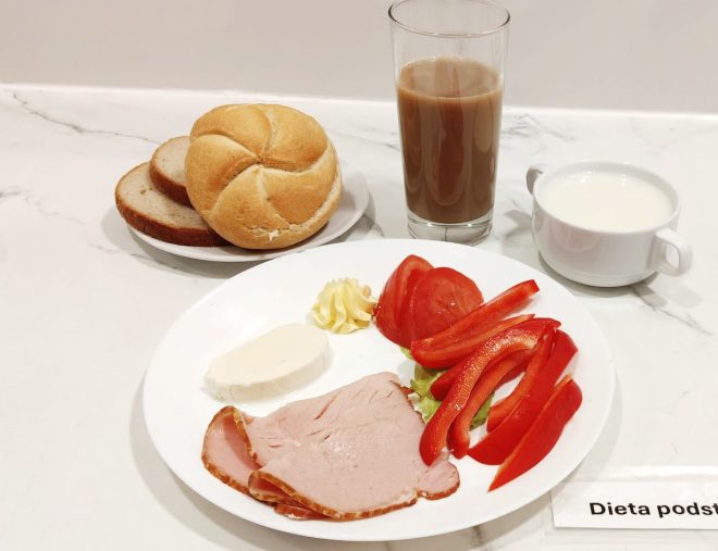 Śniadanie w diecie podstawowej. Na talerzu: plasterki czerwonej papryki, pomidor, plasterki szynki, plaster sera typu mozzarella, liść sałaty oraz niewielka porcja masła. W tle talerzyk z bułką kajzerką i kromkami chleba żytniego. Obok szklanka kawy z mlekiem oraz mała miseczka z mlekiem lub napojem mlecznym. Całość ustawiona na białym, marmurowym blacie z etykietą „Dieta podstawowa”