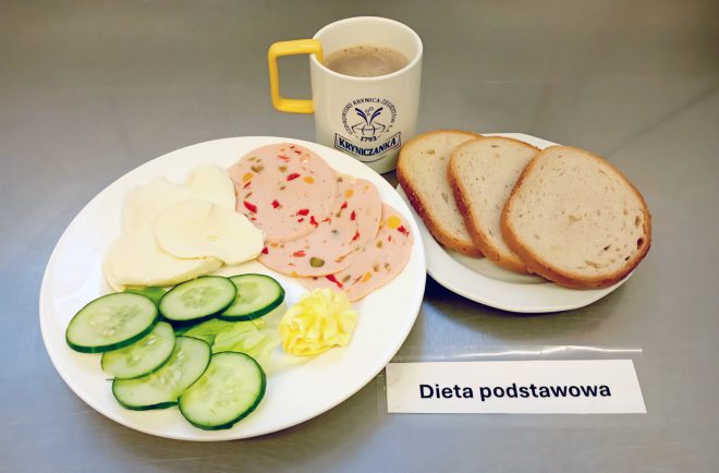 Śniadanie z „Diety podstawowej” ułożone na tacy. Na jednym talerzu trzy kromki jasnego chleba pszennego. Obok drugi talerz z dodatkami: plasterki żółtego sera, plasterki wędliny drobiowej z warzywami, ogórek świeży, liść sałaty i rozetka masła. Do zestawu dołączony kubek z napojem typu kawa zbożowa lub kawa z mlekiem – na kubku logo „Kryniczanka”