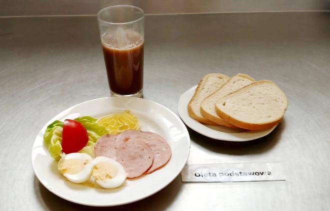 Na metalowym blacie stoi szklanka z ciemnym napojem (prawdopodobnie kawa zbożowa). Obok leży talerz z trzema kromkami białego pieczywa. Na oddzielnym talerzu znajduje się plasterek pomidora, kilka liści sałaty, dwie plasterki mielonki, pół jajka na twardo oraz kleks musztardy. Obok widoczna jest karteczka z napisem „Dieta podstawowa”