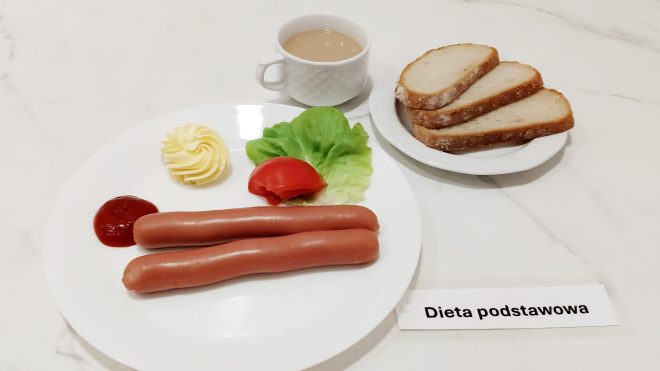 Zestaw śniadaniowy w diecie podstawowej obejmuje: dwa parzone frankfurterki podane na talerzu z dodatkiem ketchupu, masła w dekoracyjnej rozetce oraz plasterkiem pomidora i liściem sałaty, talerzyk z trzema kromkami chleba razowego z widoczną mączną posypką, filiżankę kawy z mlekiem lub kawy zbożowej z mlekiem. Posiłek ułożony jest na białym blacie, obok karteczki z napisem: „Dieta podstawowa”