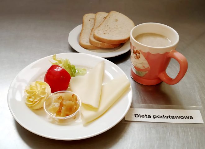 Śniadanie w ramach diety podstawowej – na talerzu znajdują się trzy plasterki sera żółtego, porcja masła uformowanego dekoracyjnie, kawałek sałaty, plaster pomidora oraz mały pojemniczek z konfiturą jabłkową. Obok kubek z kawą z mlekiem i talerzyk z czterema kromkami białego pieczywa. Na stole znajduje się etykieta z napisem „Dieta podstawowa”