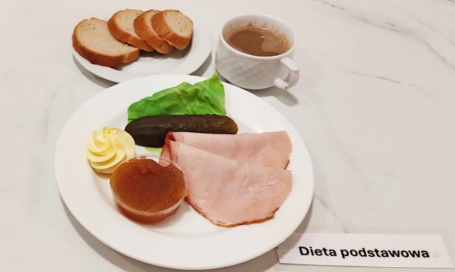 Śniadanie w diecie podstawowej na białym talerzu: dwie cienkie plastry wędliny drobiowej, liść sałaty, plaster ogórka kiszonego, rozetka masła, porcja dżemu w plastikowym pojemniczku. Obok filiżanka z kawą z mlekiem i talerzyk z kilkoma kromkami chleba pszennego. Na stole kartka z napisem „Dieta podstawowa”