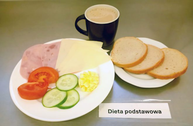 Taca z posiłkiem śniadaniowym oznaczonym jako „Dieta podstawowa”. Na jednym talerzu plastry szynki, sera żółtego, pomidora, ogórka i porcja masła. Obok drugi talerz z trzema kromkami jasnego chleba. Całość uzupełnia kubek kawy z mlekiem
