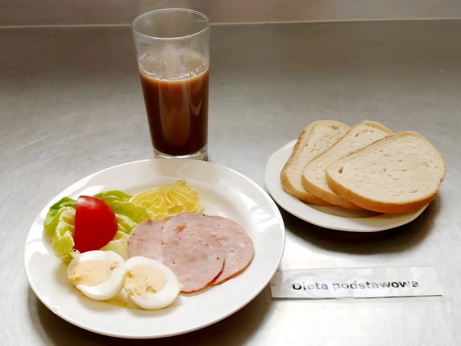 Zbliżenie na obiad z diety podstawowej: pieczone udko z kurczaka, ziemniaki z sosem i surówka z kapusty na talerzu. Obok filiżanka jasnego kompotu, miska z zupą jarzynową i gruszka. Obok leży kartka z napisem „Dieta podstawowa”
