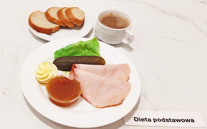 Talerz z dwoma plastrami szynki, liściem sałaty, ogórkiem konserwowym, porcją masła i galaretką owocową; obok filiżanka kawy i talerzyk z czterema kromkami chleba pszennego. Na stole karteczka z napisem „Dieta podstawowa”