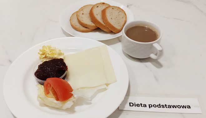 Śniadanie w diecie podstawowej na jasnym blacie. Na małym talerzu trzy kromki pieczywa mieszanego. Na większym talerzu plaster żółtego sera, porcja sałaty z plasterkiem pomidora, kostka masła oraz kubeczek z ciemnym dżemem. Obok biała filiżanka z kawą z mlekiem. W dolnym rogu widoczna karteczka z napisem „Dieta podstawowa”