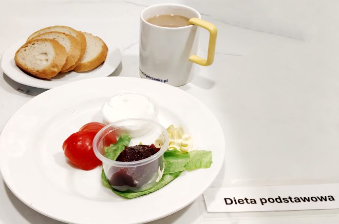 Taca z prostym śniadaniem w diecie podstawowej. Na talerzu znajdują się: dwa plasterki pomidora, liść sałaty, porcja twarożku i masła, a także porcja konfitury podana w plastikowym kubeczku. Do tego podano trzy kromki białego pieczywa oraz kubek kawy z mlekiem w białym kubku z żółtym uchem. Zestaw jest oznaczony etykietą „Dieta podstawowa”