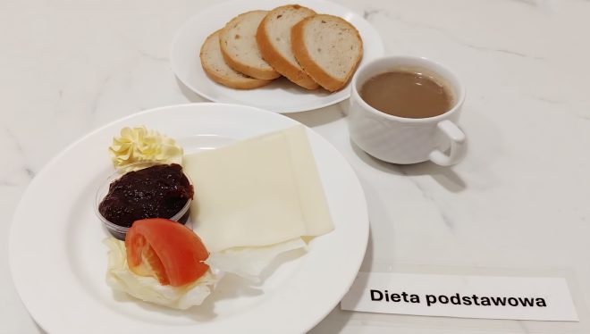Talerz śniadaniowy z plastrem sera żółtego, porcją masła, konfiturą, plastrem pomidora i liściem sałaty. W tle osobny talerzyk z trzema kromkami chleba oraz filiżanka kawy z mlekiem. Na blacie widoczna etykieta z napisem „Dieta podstawowa”