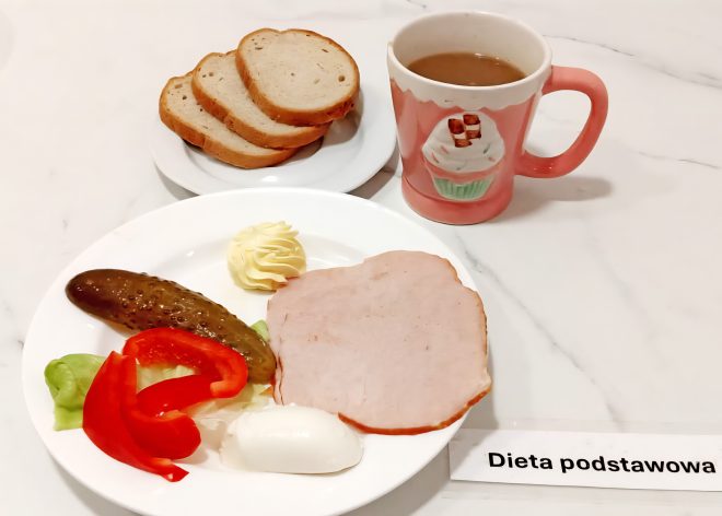Białe pieczywo pszenne w towarzystwie gotowanego jajka, plasterka szynki, ogórka konserwowego, liścia sałaty i czerwonej papryki. Na talerzu również porcja masła. Obok kubek z kawą z mlekiem. Danie podane na białym talerzu i podpisane jako „Dieta podstawowa”
