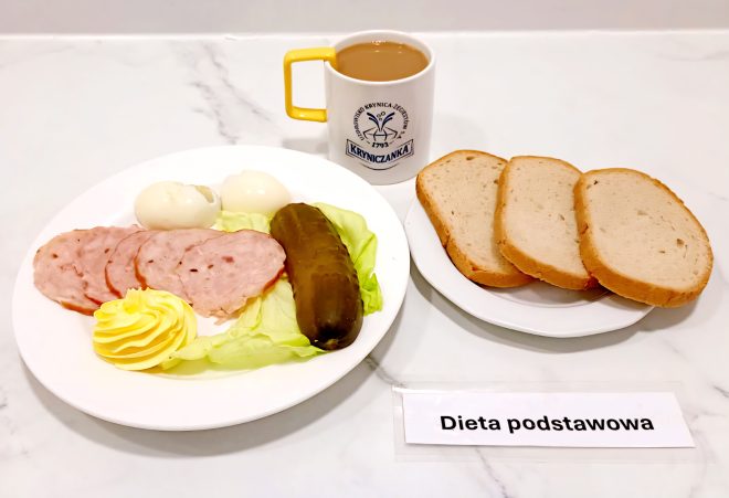 Taca ze śniadaniem szpitalnym w ramach „Diety podstawowej”. Na dużym białym talerzu znajdują się dwa jajka na twardo, trzy plasterki mielonki drobiowej, liść sałaty, ogórek konserwowy oraz porcja masła w kształcie rozetki. Obok osobny talerzyk z trzema kromkami białego pieczywa. Do śniadania podano kawę z mlekiem w białym kubku z żółtym uchem i logo „Kryniczanka”