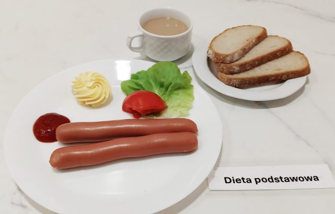 Śniadanie na diecie podstawowej. Na talerzu dwie parówki z ketchupem i rozetką z masła, obok liść sałaty i kawałek pomidora. Na osobnym talerzyku trzy kromki jasnego chleba. Do tego kubek z kawą z mlekiem. Na stole etykieta z napisem "Dieta podstawowa"