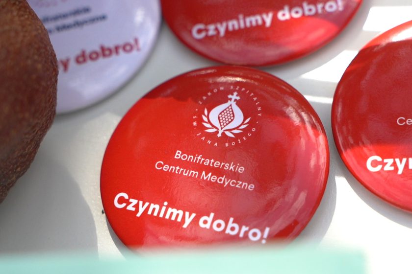 Przypinki z hasłem „Czynimy dobro!” – Zbliżenie na kolorowe przypinki z logo Zakonu Bonifratrów i napisem „Czynimy dobro!”, ułożone na stole obok owoców