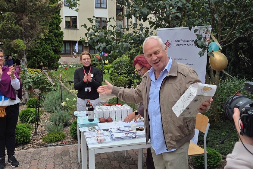 Mężczyzna trzymający upominek – Uśmiechnięty mężczyzna prezentuje otrzymany upominek podczas spotkania w ogrodzie Bonifraterskiego Centrum Medycznego. W tle stół z materiałami informacyjnymi i pracownicy centrum