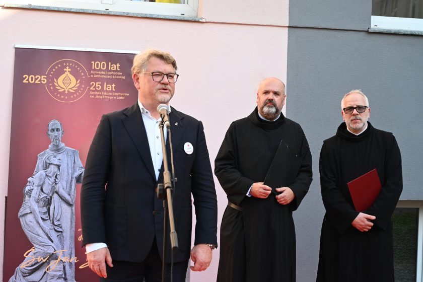 Mężczyzna w garniturze przemawia przy mikrofonie podczas uroczystości jubileuszowych przed banerem Zakonu Bonifratrów. Obok niego stoją dwaj zakonnicy w czarnych habitach