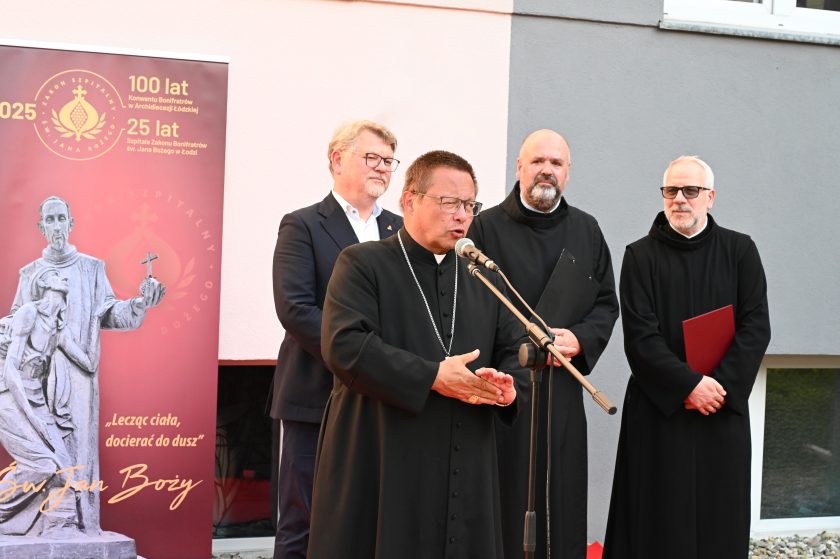 Biskup w czarnej sutannie przemawia przy mikrofonie podczas uroczystości jubileuszowej Bonifratrów. Za nim stoją zakonnicy w habitach i mężczyzna w garniturze