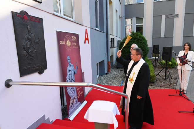 Biskup święci odsłoniętą tablicę jubileuszową Bonifratrów, stojąc przy niej z kropidłem. Obok zakonnicy i prowadząca wydarzenie
