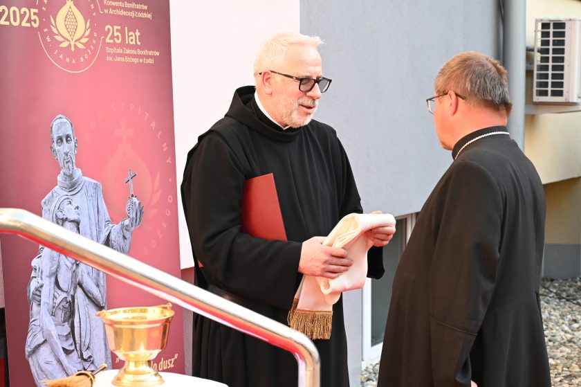 Rozmowa zakonnika z biskupem podczas uroczystości jubileuszowych, w tle baner z wizerunkiem św. Jana Bożego